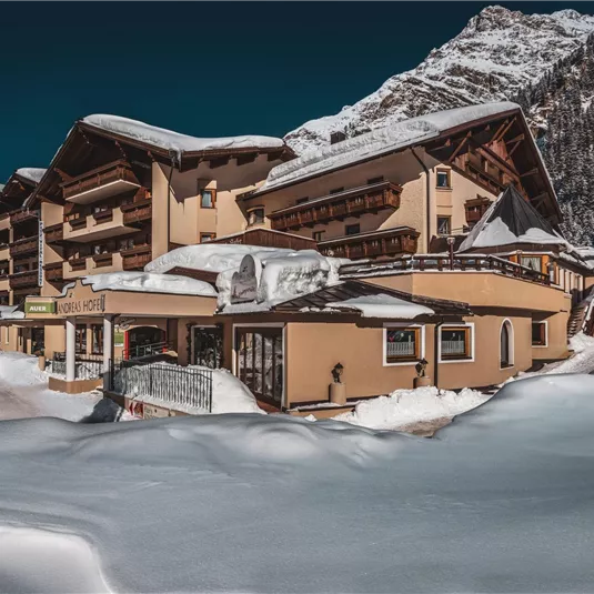 Ein gemütliches Hotel in den Alpen, umgeben von schneebedeckten Bergen. Der Himmel ist klar und die Landschaft bietet eine winterliche Idylle.