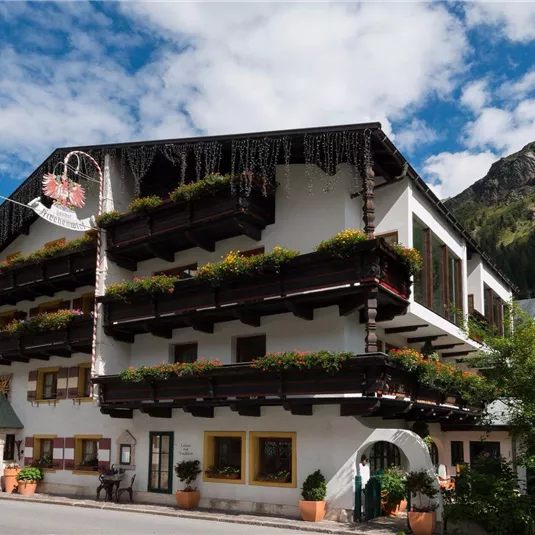 Ein gemütliches Hotel mit traditioneller Architektur und vielen Blumenbalkonen. Im Hintergrund sind Berge und ein blauer Himmel zu sehen.