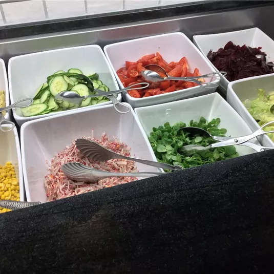 Eine Auswahl an frischen Salatzutaten in weißen Schalen. Zu sehen sind Gurken, Tomaten, Mais, rote Beete, Salat und geschnittenes Fleisch.