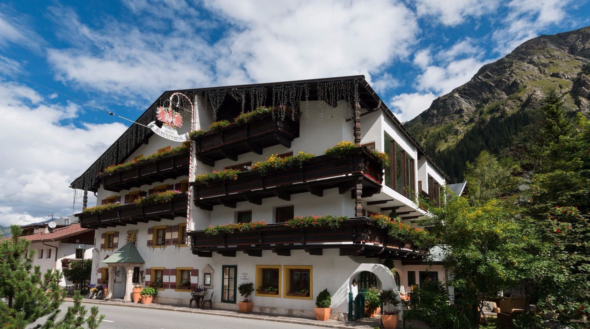Ein gemütliches Hotel mit traditioneller Architektur und vielen Blumenbalkonen. Im Hintergrund sind Berge und ein blauer Himmel zu sehen.