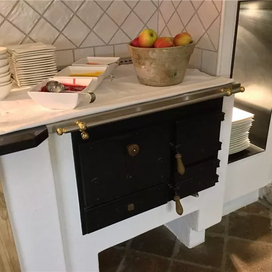 Eine alte Küche mit einem schwarz lackierten Ofen und einer Arbeitsplatte. Oben befinden sich Äpfel in einer Schüssel und Teller im Hintergrund.