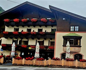 A traditional hotel with blooming balcony boxes and an inviting terrace. The surrounding mountains are visible, creating a picturesque atmosphere.