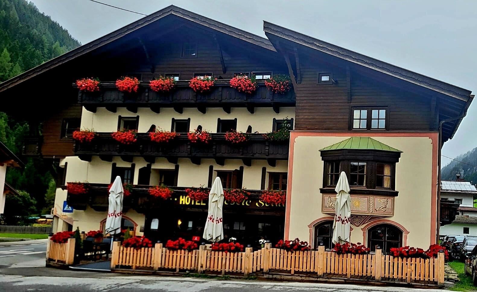 A traditional hotel with blooming balcony boxes and an inviting terrace. The surrounding mountains are visible, creating a picturesque atmosphere.