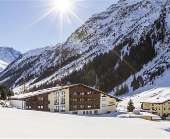 Eine malerische Gebirgslandschaft mit schneebedeckten Bergen. Im Vordergrund befindet sich ein gemütliches Hotel, umgeben von weißem Schnee.