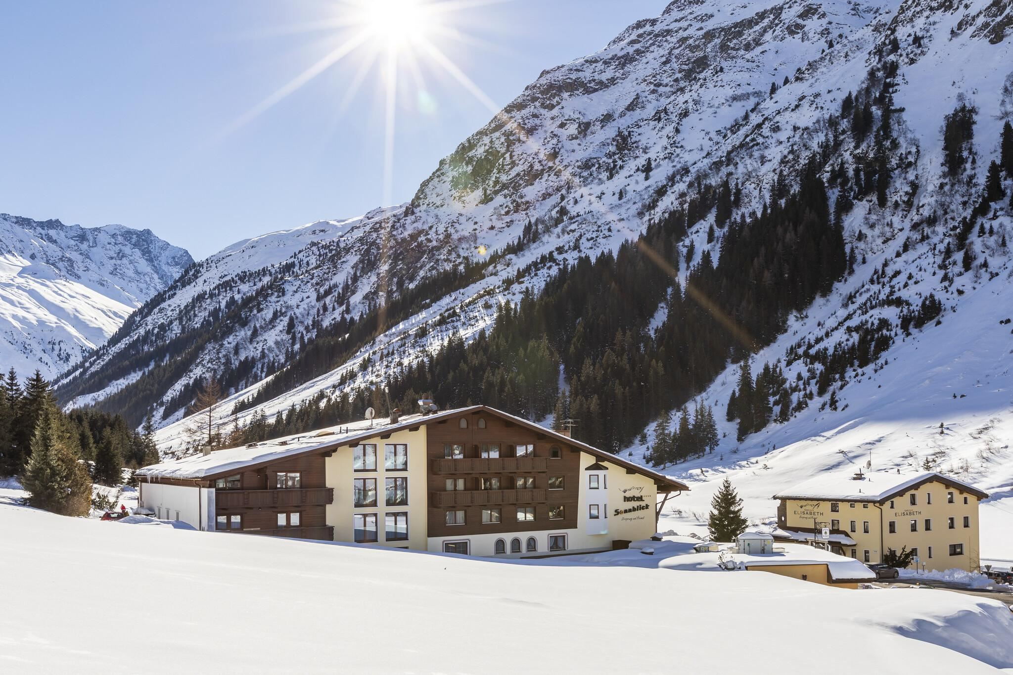 Eine malerische Gebirgslandschaft mit schneebedeckten Bergen. Im Vordergrund befindet sich ein gemütliches Hotel, umgeben von weißem Schnee.