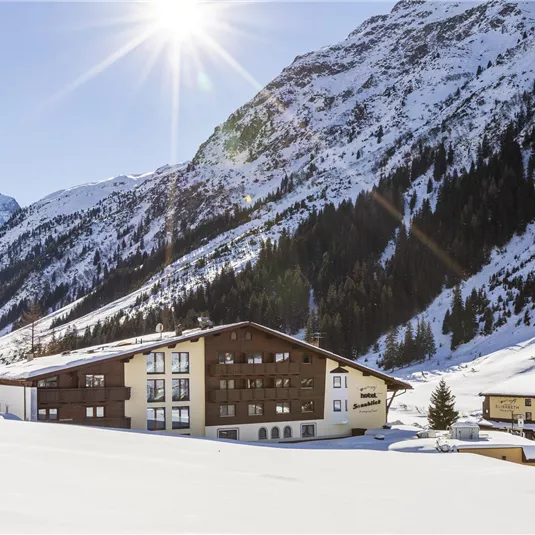 Eine malerische Gebirgslandschaft mit schneebedeckten Bergen. Im Vordergrund befindet sich ein gemütliches Hotel, umgeben von weißem Schnee.