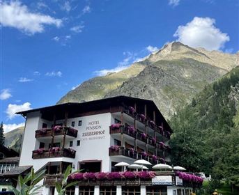 Ein charmantes Hotel mit Balkonen voller Blumen, umgeben von majestätischen Bergen. Der Himmel ist blau mit einigen Wolken.