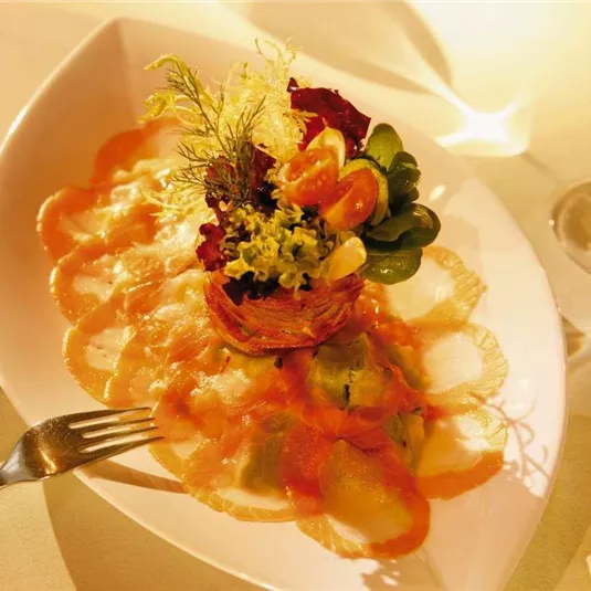Ein kunstvoll angerichteter Teller mit Fisch-Carpaccio und buntem Salat. Im Hintergrund ist ein Glas Wein zu sehen.