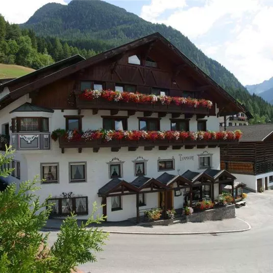 Ein charmantes Hotel in einer bergigen Landschaft, umgeben von Bäumen und grünen Wiesen. Die Fassade ist mit Blumen dekoriert und vermittelt eine einladende Atmosphäre.