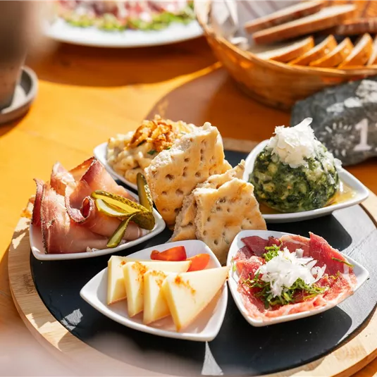 Ein Teller mit verschiedenen Vorspeisen, darunter Wurst, Käse, Crackers und eine grüne Spinatmasse. Die Speisen sind schön angerichtet und auf einem Holzbrett serviert.