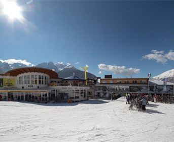 Ein Skigebiet mit viel Schnee und einem modernen Gebäude im Vordergrund. Im Hintergrund sind Berge und blauer Himmel zu sehen.