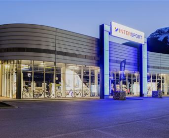 A modern store from INTERSPORT with large windows and a illuminated entrance. Mountains are visible in the background, and the evening dusk creates a moody atmosphere.