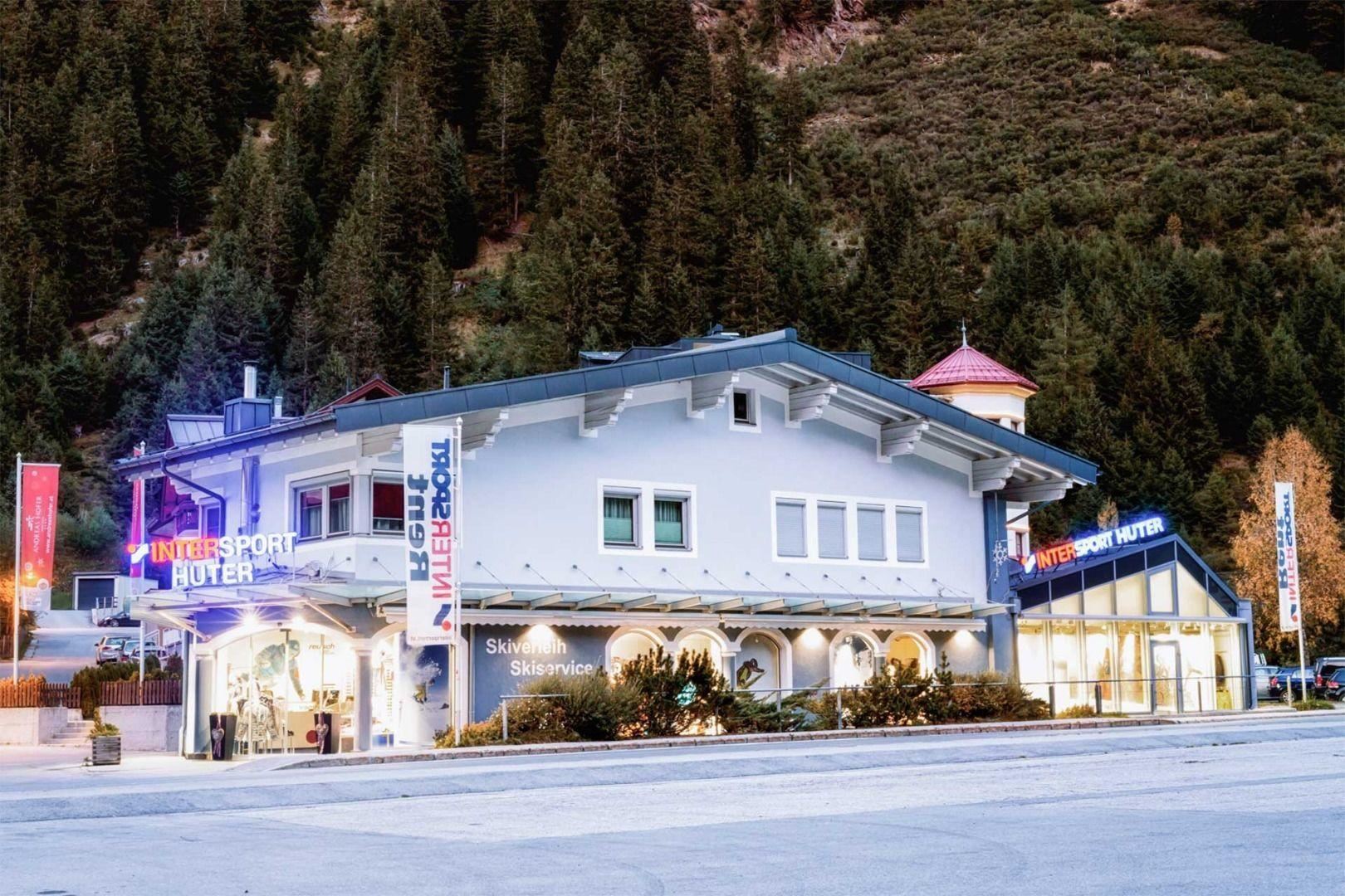 Ein modernes Skigeschäft mit auffälligen Schildern in den Alpen. Umgeben von Bäumen und Bergen bietet es Equipment und Dienstleistungen für Wintersportler an.