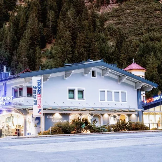 Ein modernes Skigeschäft mit auffälligen Schildern in den Alpen. Umgeben von Bäumen und Bergen bietet es Equipment und Dienstleistungen für Wintersportler an.