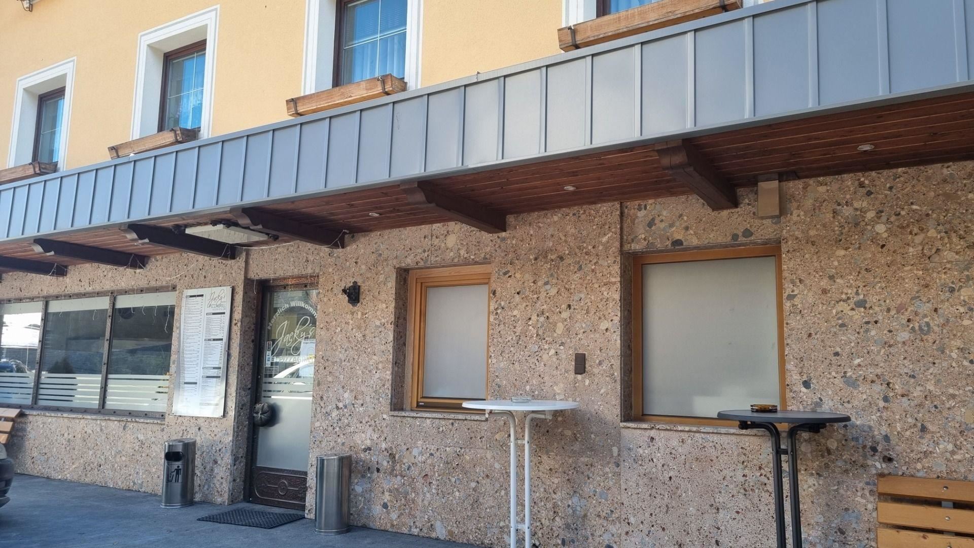 Ein Gebäude mit einer Steinfassade und Holzveranda. Die Fenster sind teilweise geschlossen, und es gibt Tische im Außenbereich.