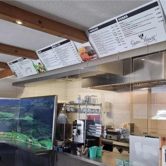 Ein gemütliches Restaurantinnere mit einem großen Menü an der Wand. Im Hintergrund ist eine digitale Anzeige mit Landschaftsbildern zu sehen.