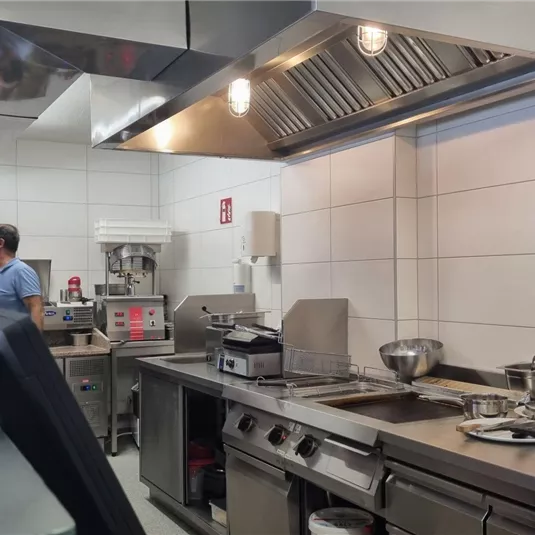 Eine moderne Küche mit Edelstahlgeräten und einer sauberen Arbeitsfläche. Im Hintergrund ist eine Person mit Kochutensilien beschäftigt.