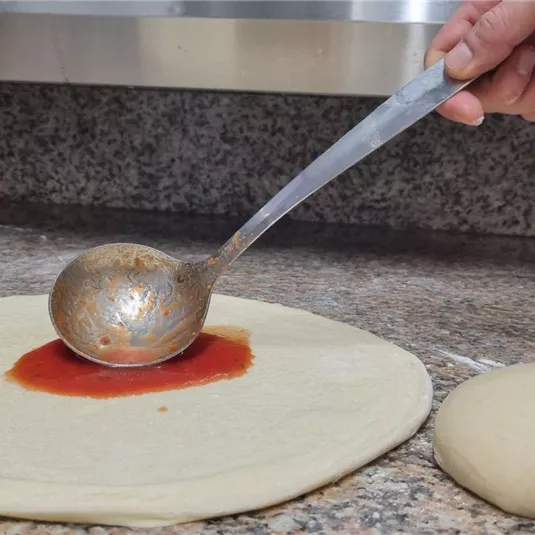 Eine Hand verteilt Tomatensauce auf einem flachen Teig. Daneben liegt eine Portion Teig auf einer Arbeitsfläche.