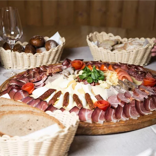 A delicious cheese and sausage platter with fresh herbs and tomatoes. In the background, there are baskets with bread and a bottle of wine.