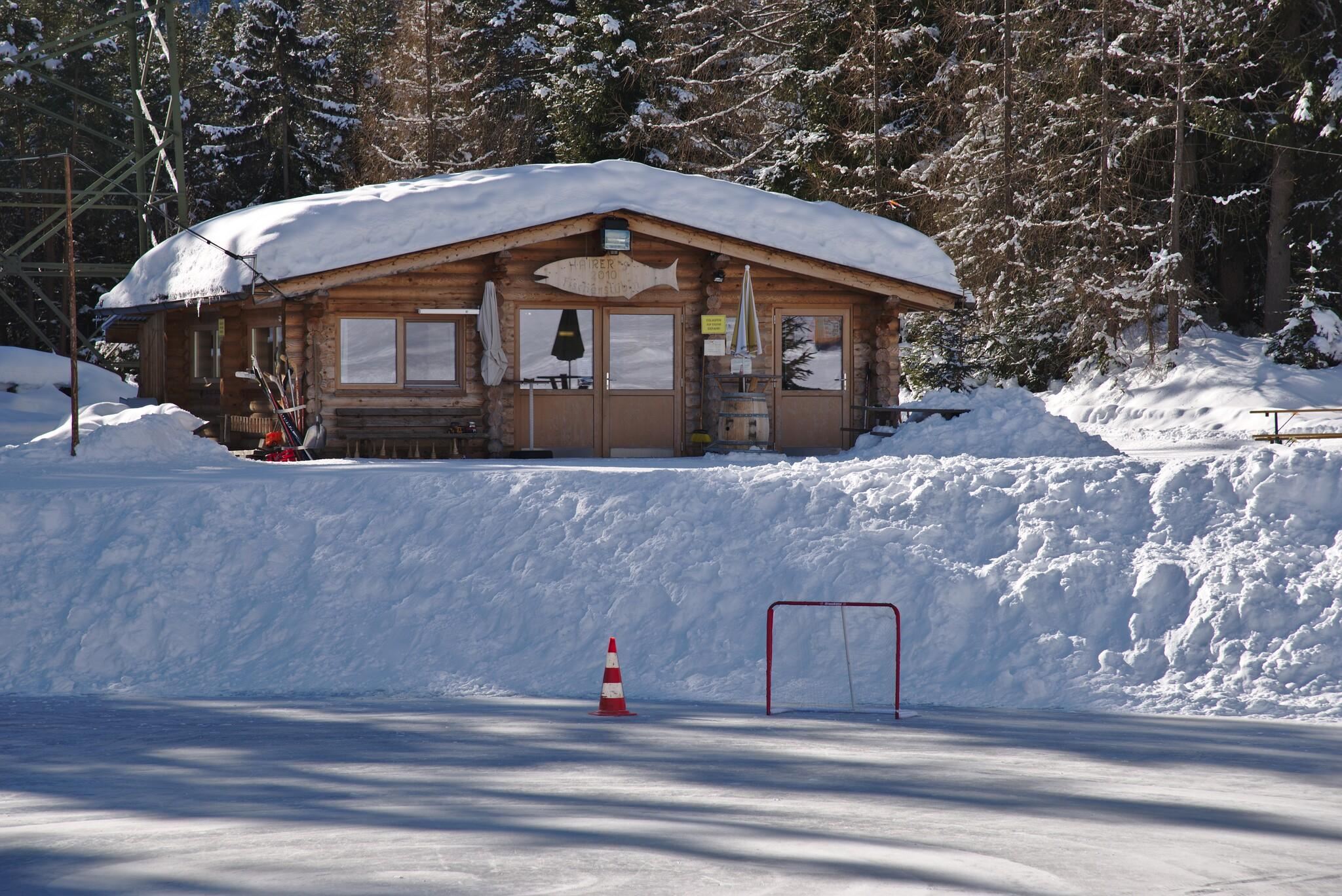Eine gemütliche Hütte im Schnee umgeben von Bäumen. Im Vordergrund befindet sich ein kleiner Eishockey-Tor.