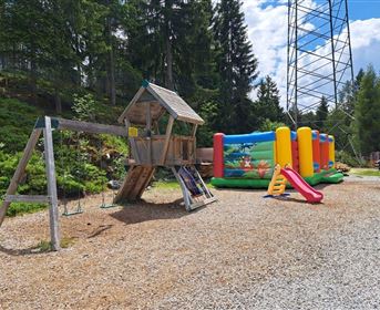 Ein Spielplatz mit einer Holzspielstruktur und einer Rutsche sowie einer aufblasbaren Hüpfburg in bunten Farben. Umgeben von Bäumen und einer klaren Blauen Himmel.