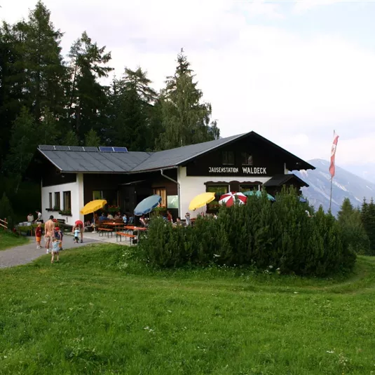 A cozy mountain restaurant surrounded by green meadows. Colorful sun umbrellas stand on the terrace and visitors enjoy the view.