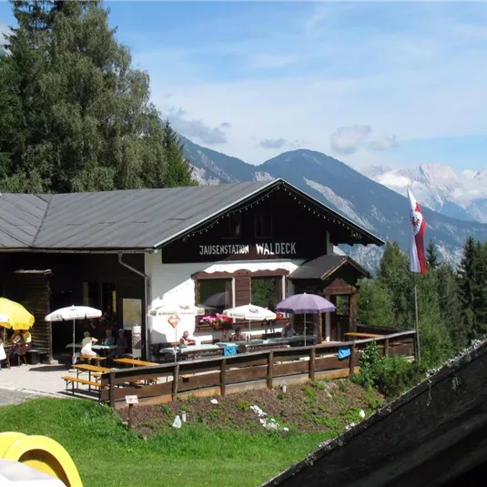 A cozy mountain cabin with a terrace and sun umbrellas. In the background, majestic mountains and forest can be seen.
