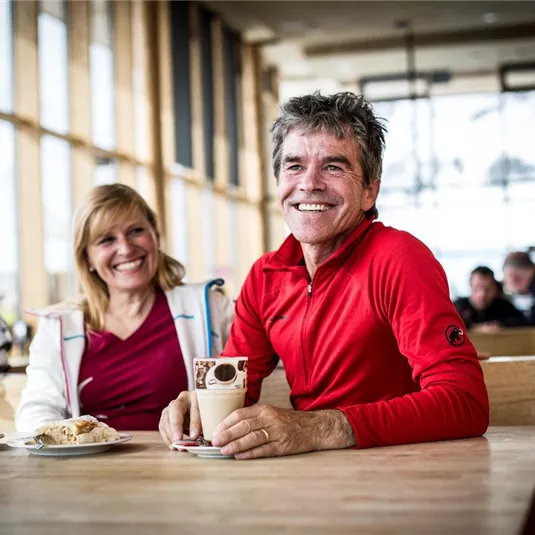 Ein fröhliches Paar sitzt an einem Tisch in einer gemütlichen Hütte. Sie genießen heiße Getränke und haben eine entspannte Zeit.