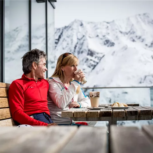 Ein Paar sitzt entspannt an einem Tisch auf einer Terrasse mit Blick auf schneebedeckte Berge. Die Frau genießt ein Getränk, während der Mann sie anlächelt.