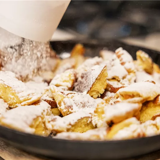 Ein Teller mit frisch zubereitetem Kaiserschmarrn, der mit Puderzucker bestreut wird. Die goldbraunen Stücke fallen verführerisch auseinander.