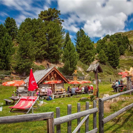 Eine gemütliche Almhütte umgeben von Bäumen und Wiesen. Menschen sitzen unter roten Sonnenschirmen und genießen die Natur.