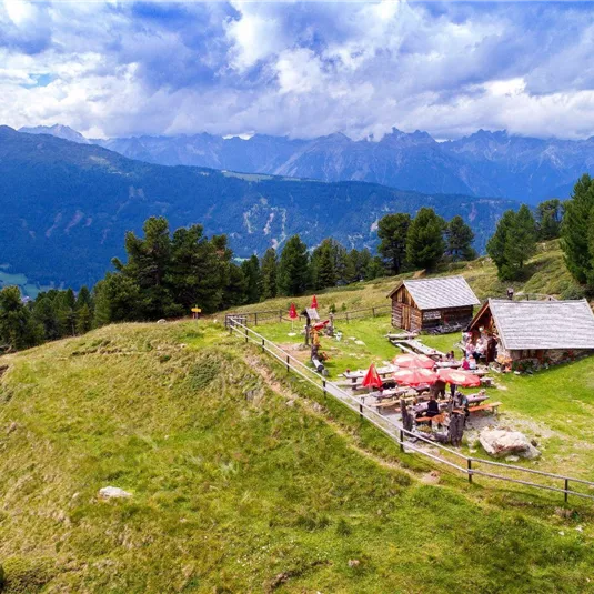 Eine idyllische Berglandschaft mit grünen Wiesen und majestätischen Bergen im Hintergrund. Im Vordergrund befinden sich gemütliche Hütten und ein kleiner Bereich mit Tischen und Sonnenschirmen.