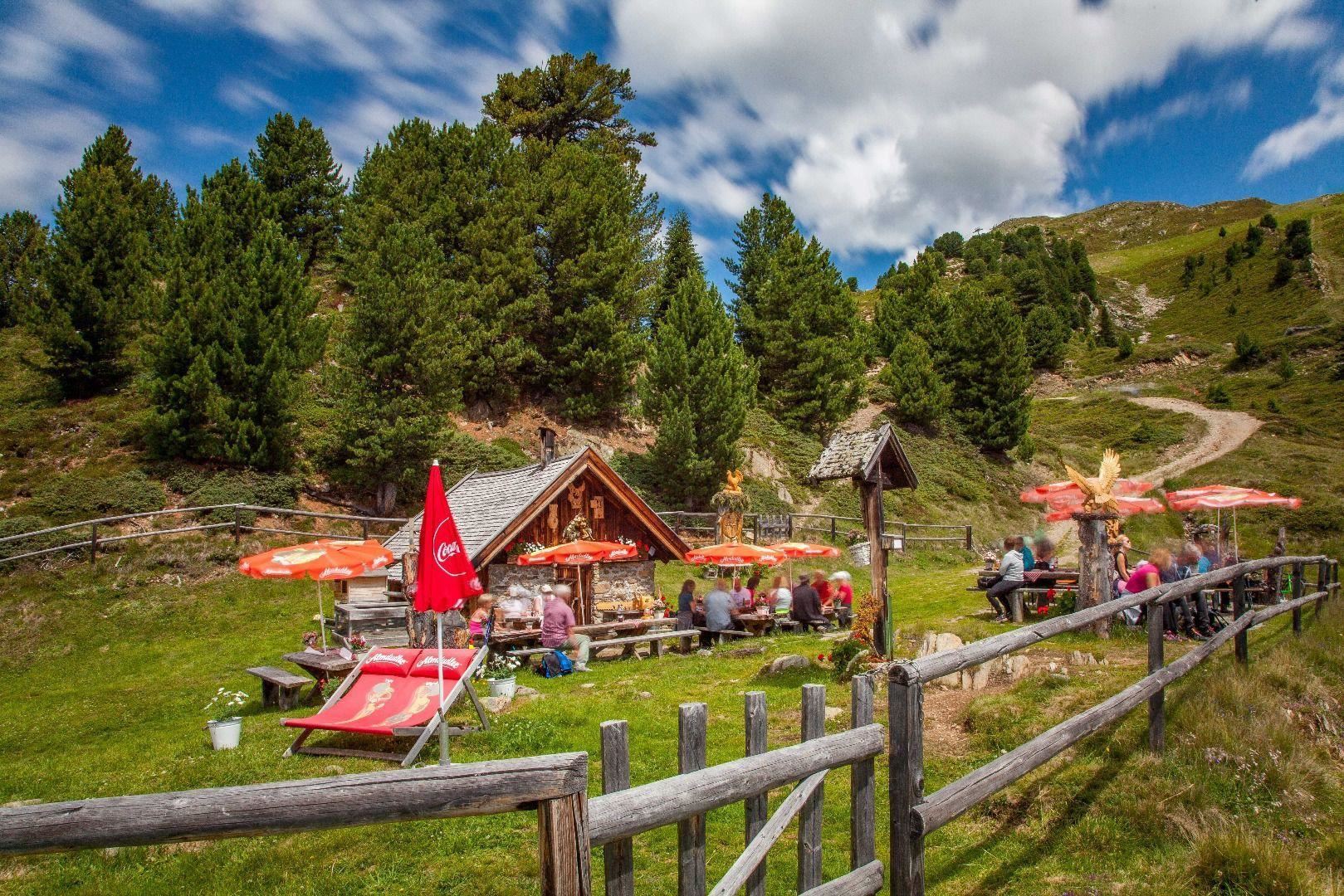 Eine gemütliche Almhütte umgeben von Bäumen und Wiesen. Menschen sitzen unter roten Sonnenschirmen und genießen die Natur.