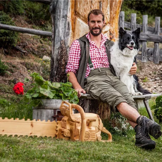 Ein Mann sitzt fröhlich auf einem Baumstumpf mit einem Hund neben sich. Im Vordergrund sind eine Holzsäge und rote Geranien zu sehen.