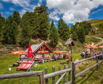 Ein gemütliches Gebirgsrestaurant im Freien mit Gästen, die sich unter roten Sonnenschirmen entspannen. Umgeben von grünen Bäumen und einer malerischen Landschaft.