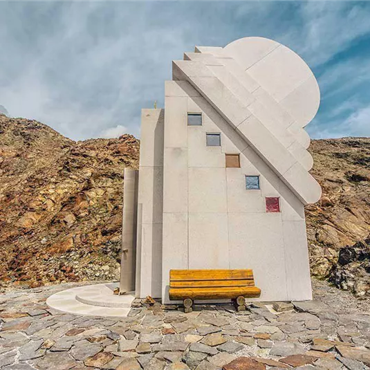 A modern architectural building stands in a rocky landscape. In front of the building, there is a wooden bench.