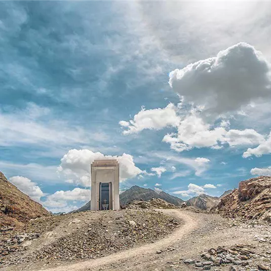 Eine verlassene Struktur auf einem felsigen Weg, umgeben von Bergen und Wolken. Der Himmel ist hell und klar.