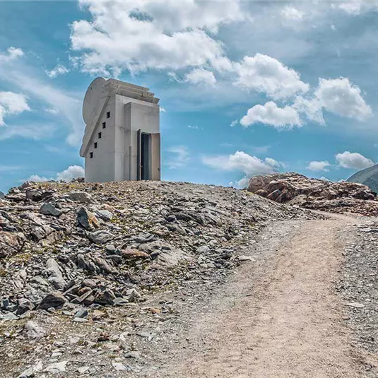 Ein modernes Gebäude auf einem felsigen Hügel, umgeben von Berglandschaft. Der Himmel ist klar mit einigen Wolken.