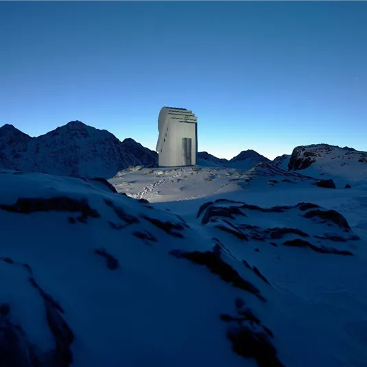 Ein modernes Gebäude steht allein in einer schneebedeckten Berglandschaft. Der Himmel ist klar und vermittelt eine ruhige, kalte Atmosphäre.