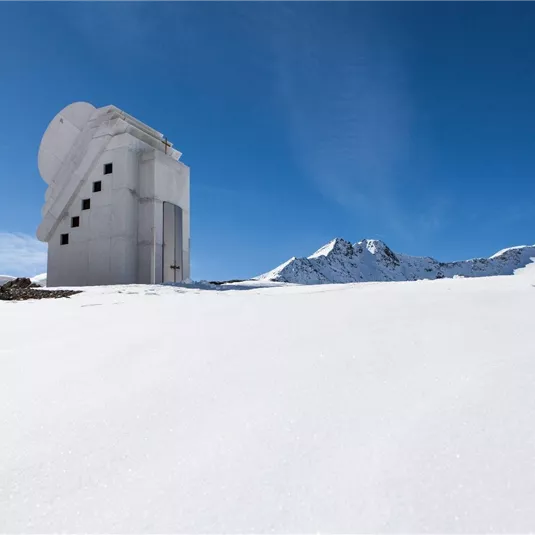 Ein modernes Gebäude steht in einer schneebedeckten Landschaft unter einem klaren blauen Himmel. Die umliegenden Berge sind teilweise mit Schnee bedeckt.