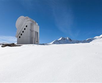 Ein modernes Gebäude steht in einer schneebedeckten Landschaft unter einem klaren blauen Himmel. Die umliegenden Berge sind teilweise mit Schnee bedeckt.
