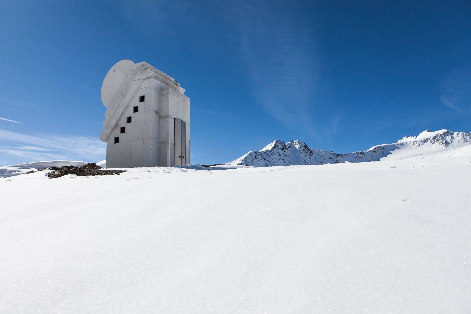 Ein modernes Gebäude steht in einer schneebedeckten Landschaft unter einem klaren blauen Himmel. Die umliegenden Berge sind teilweise mit Schnee bedeckt.