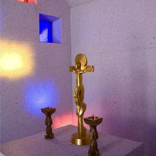 Eine goldene Skulptur auf einem Steinaltar, umgeben von zwei kleinen Kerzenhaltern. Farbige Lichtakzente erhellen den Raum.