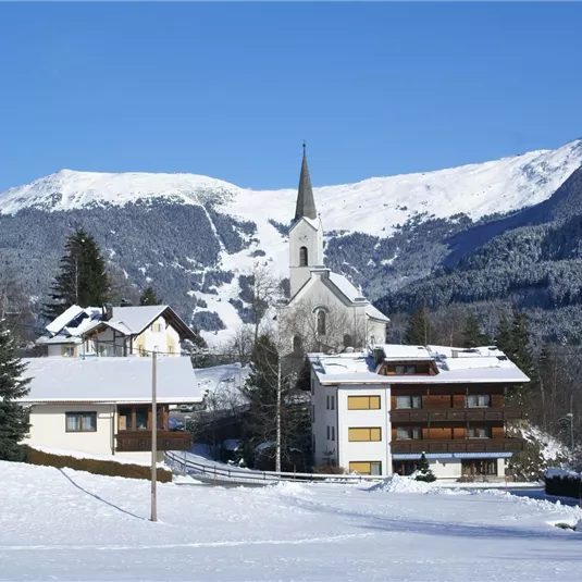 Eine winterliche Landschaft mit schneebedeckten Häusern und einer Kirche im Vordergrund. Im Hintergrund sind majestätische Berge und ein blauer Himmel zu sehen.