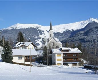 Eine winterliche Landschaft mit schneebedeckten Häusern und einer Kirche im Vordergrund. Im Hintergrund sind majestätische Berge und ein blauer Himmel zu sehen.
