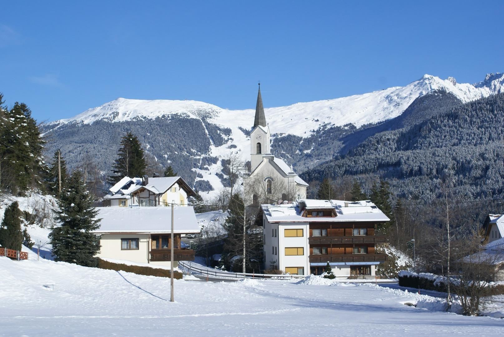 Eine winterliche Landschaft mit schneebedeckten Häusern und einer Kirche im Vordergrund. Im Hintergrund sind majestätische Berge und ein blauer Himmel zu sehen.