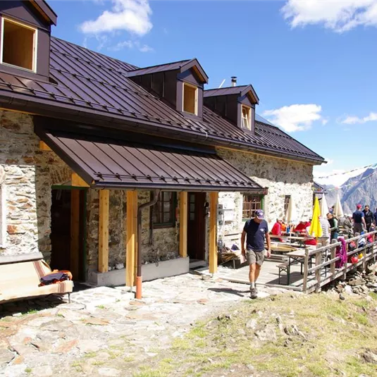 Ein traditionelles Berghaus in den Alpen mit einer schönen Terrasse. Im Hintergrund sind beeindruckende Berge und ein klarer Himmel zu sehen.