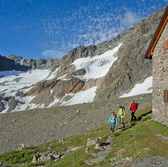 Eine schöne Berglandschaft mit schneebedeckten Gipfeln. Wanderer gehen an einer gemütlichen Hütte vorbei.