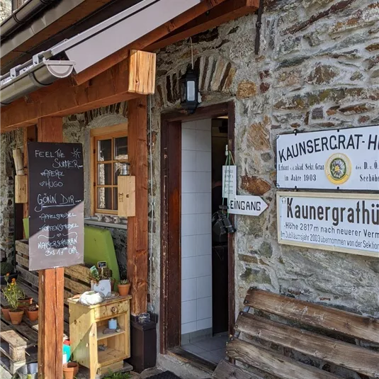 Ein rustikales Gebäude aus Stein mit einem einladenden Eingang. Vor der Tür steht eine Holzbank und es gibt ein Schild mit Informationen.