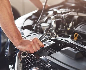 A mechanic is working on a vehicle engine. In his hand, he holds a wrench.